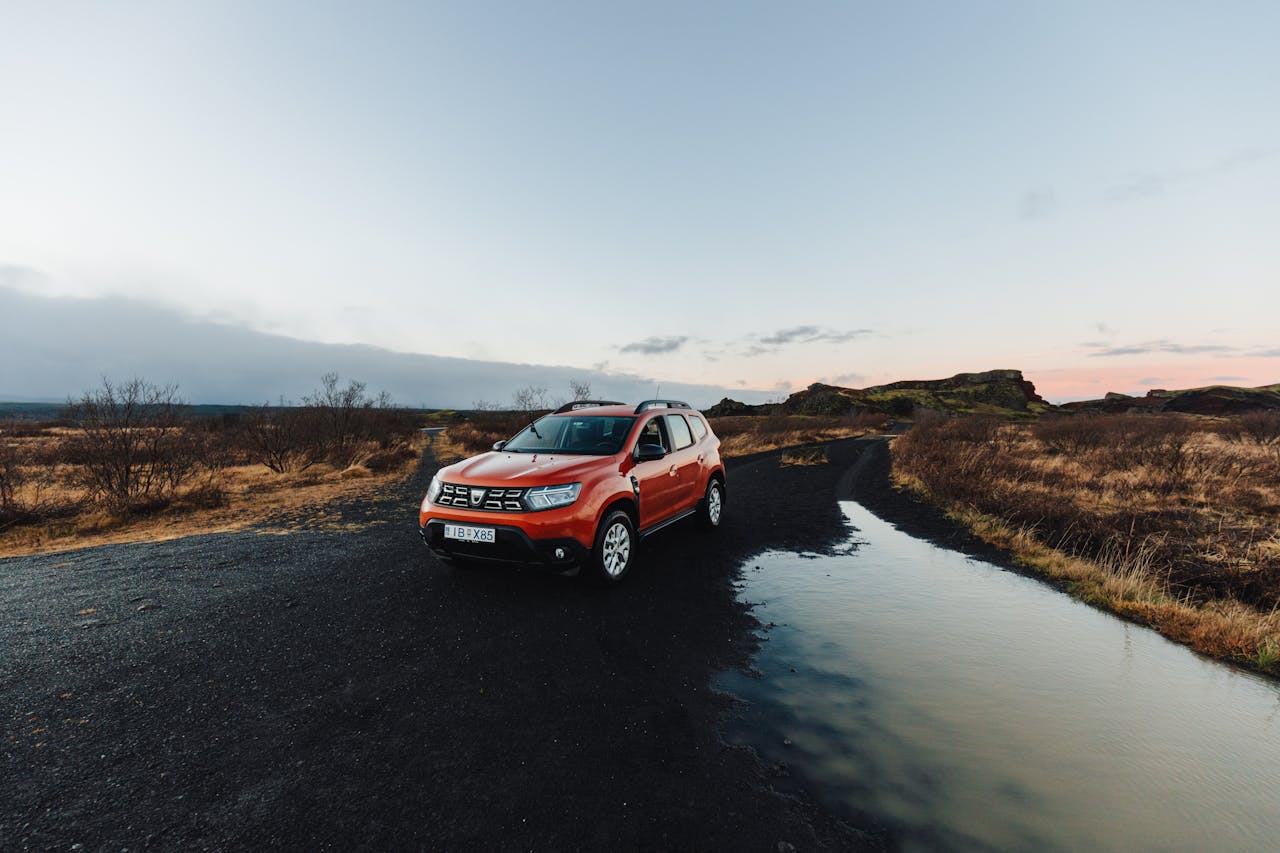A red SUV parked on a rural dirt road with a puddle, under a clear sky at sunset.